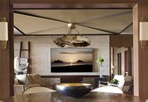 A modern living room with a large flat-screen TV mounted on a textured wall, a wooden dining table with chairs, and a unique hanging light fixture.
