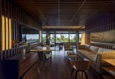 A modern interior with a wooden slatted ceiling, large windows offering a view of palm trees, and a seating area with a sofa and chairs.