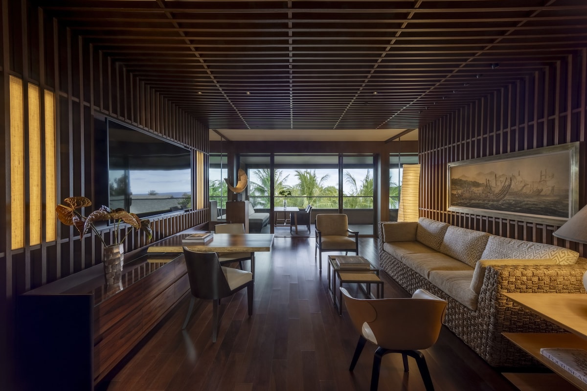 A modern interior with a wooden slatted ceiling, large windows offering a view of palm trees, and a seating area with a sofa and chairs.
