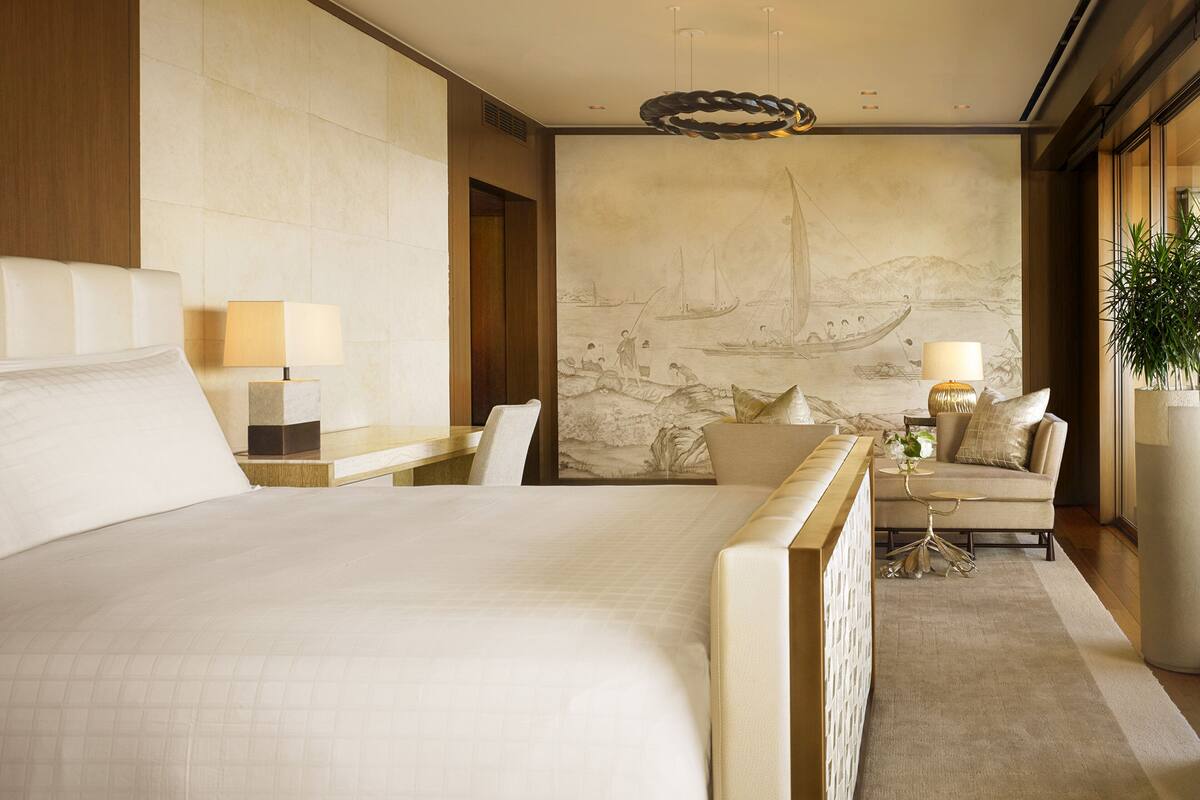 Ein Hotelzimmer mit einem großen Bett, einer Sitzecke mit Sofa und Sesseln, einem Esstisch und einem Wandgemälde.