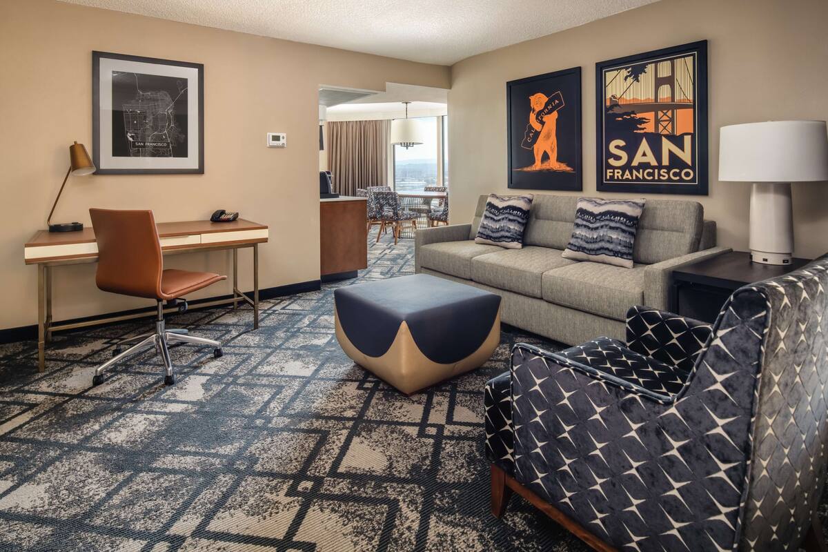 Een hotelkamer met een bureau, een bank, een getextureerd vloerkleed en kunstwerken die San Francisco voorstellen.