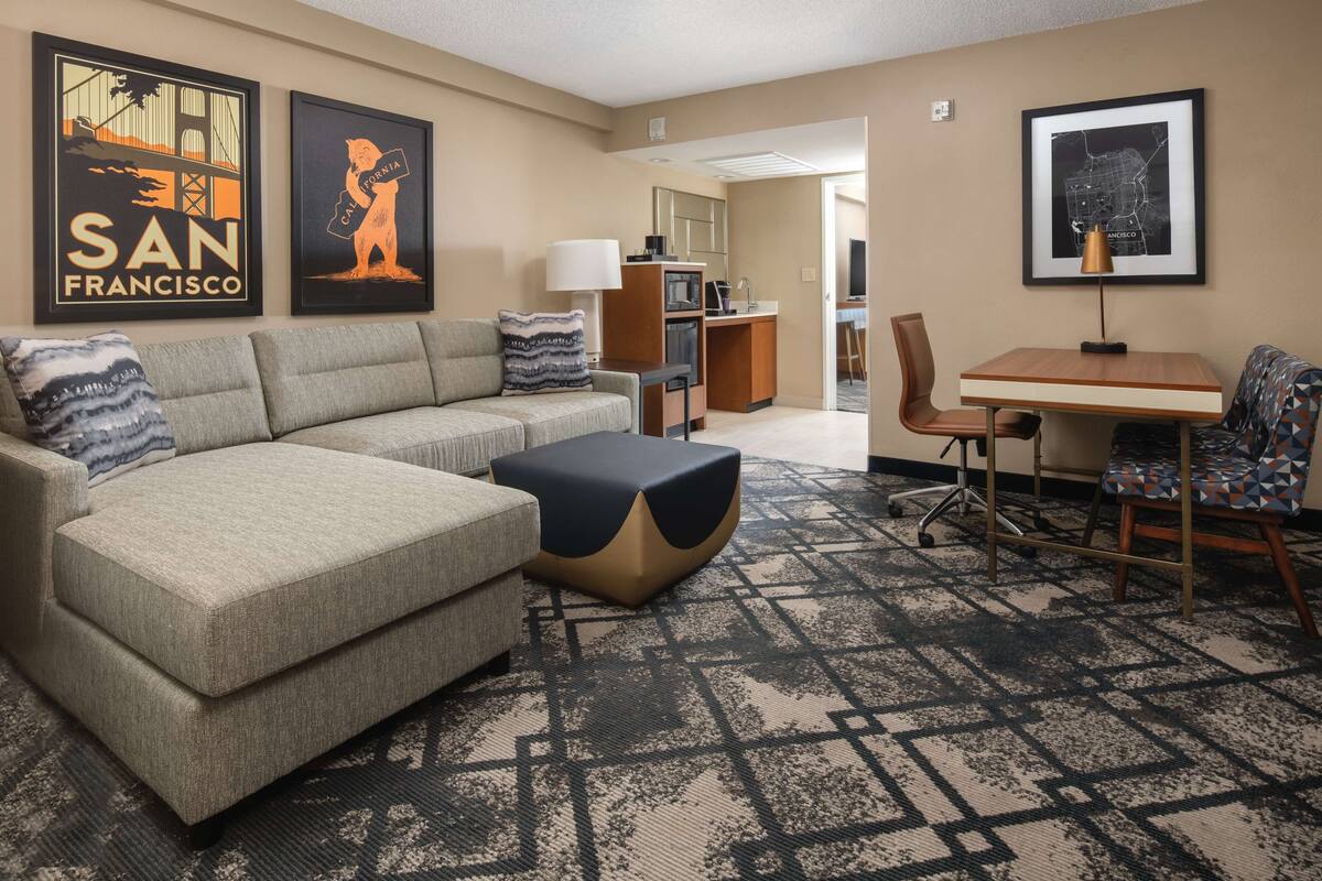Een hotelkamer met een bank, een salontafel, een bureau en stoelen. Er hangen afbeeldingen in lijsten aan de muur en de vloer is voorzien van tapijt.