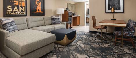 A hotel room with a sofa, a coffee table, a desk, and chairs. There are framed pictures on the wall and a carpeted floor.