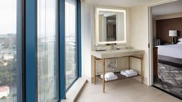 A modern hotel bathroom with a large window, a sink with a mirror, and a bed with a patterned headboard.