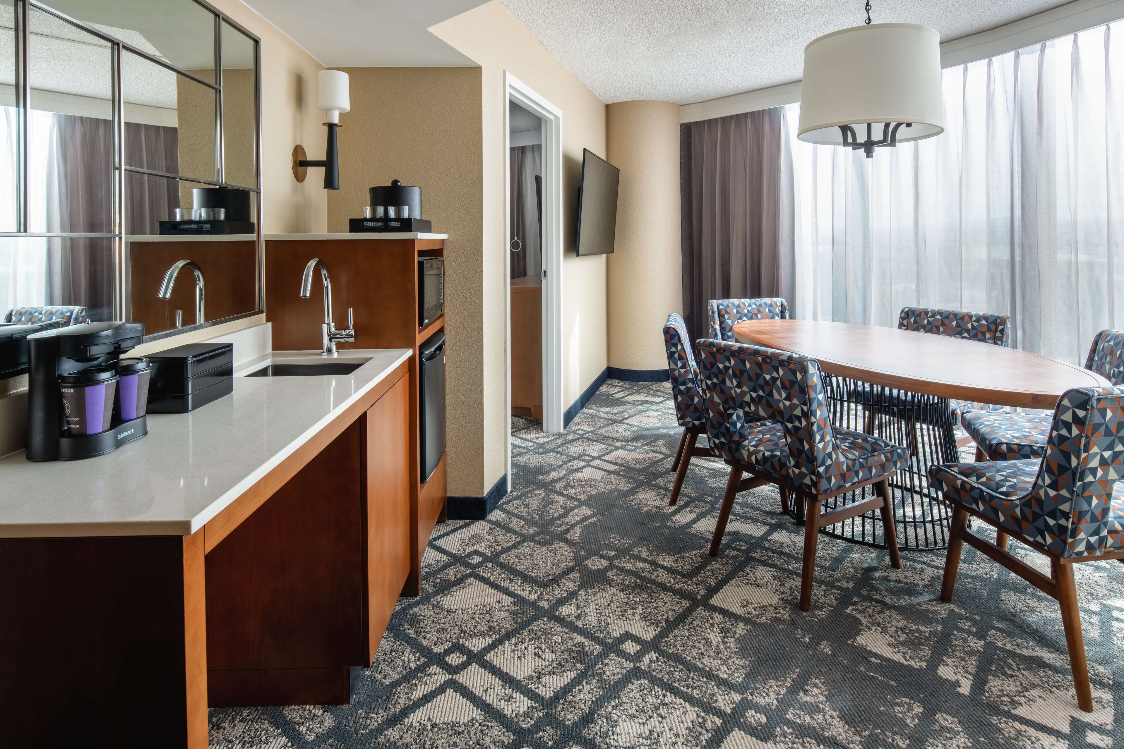 A hotel room with a kitchenette, a dining area with patterned chairs, a large mirror, and a TV.