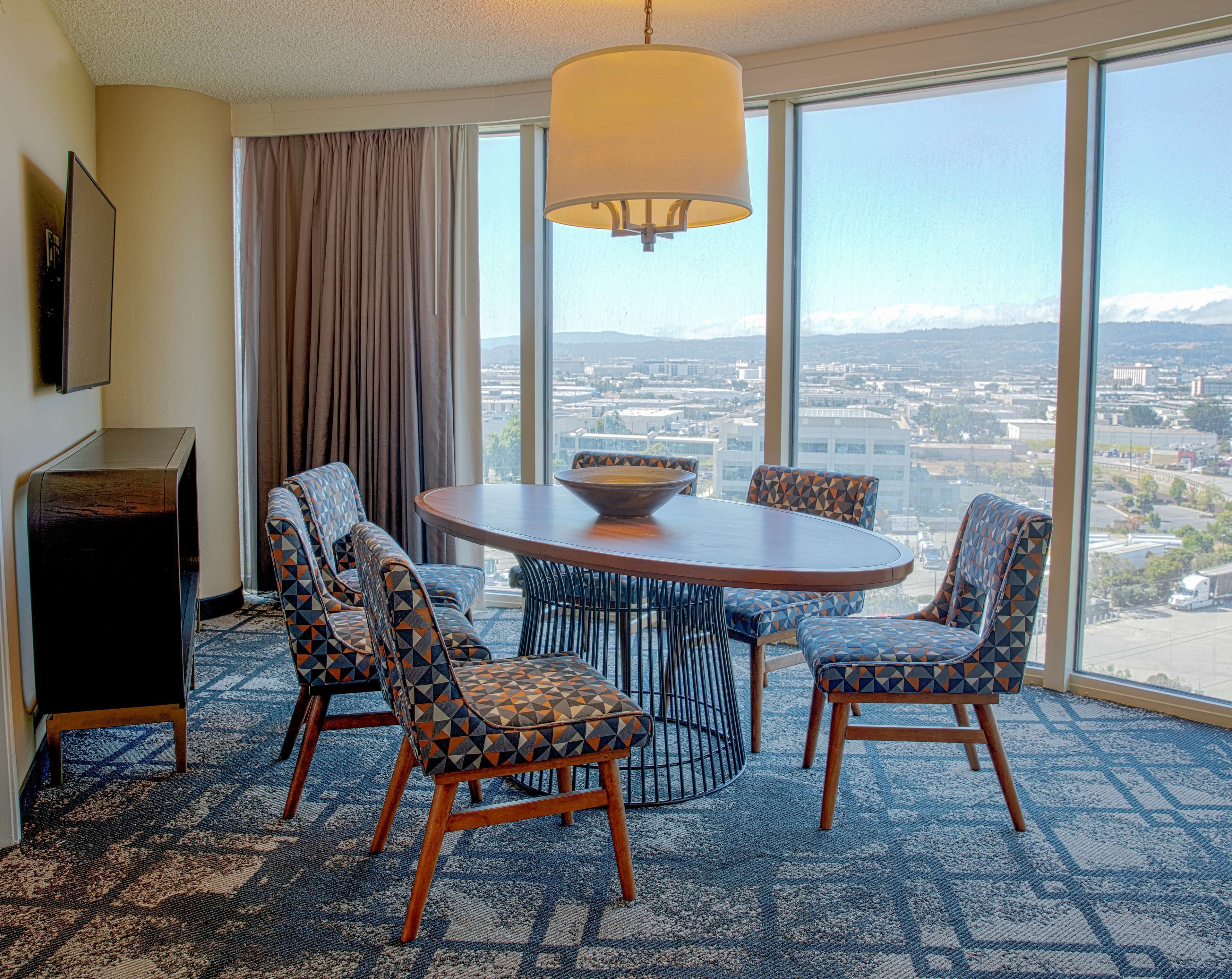 Une chambre d’hôtel avec une grande fenêtre, une table ronde entourée de chaises, une lampe et un téléviseur posé sur un meuble.