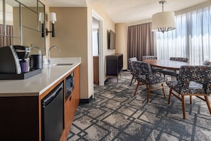 A hotel room with a kitchenette, a dining area with patterned chairs, a large mirror, and a chandelier.