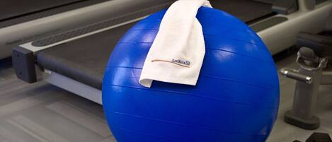 A blue exercise ball with a white towel on top, placed on a gym floor with a treadmill and dumbbells nearby.