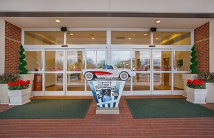 Entrada de un edificio con puertas dobles, una escultura decorativa de un coche y plantas en macetas.