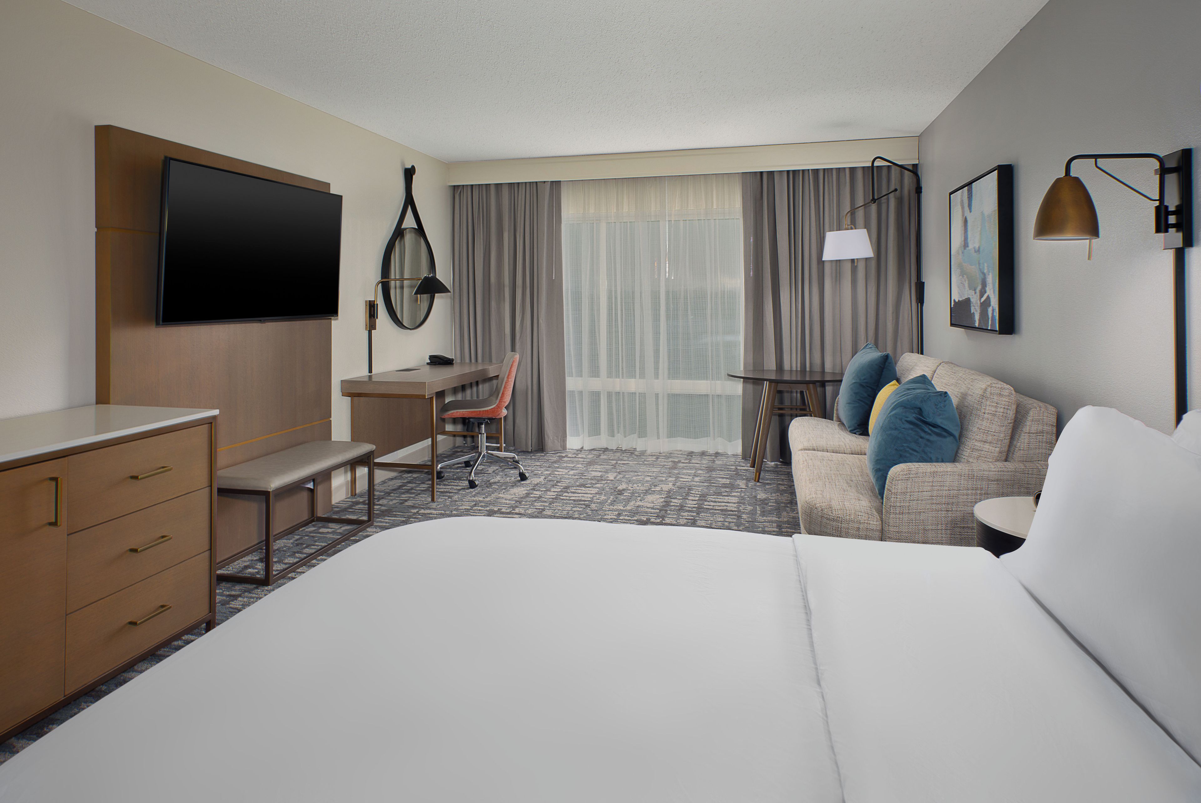 A hotel room with a bed, a desk, a chair, a TV, a lamp, and a window with curtains.