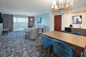 A hotel room with a large dining table, blue chairs, a sofa, a TV, and a window with curtains. - Crowne Plaza Bowling Green KY by IHG (Bowling Green)