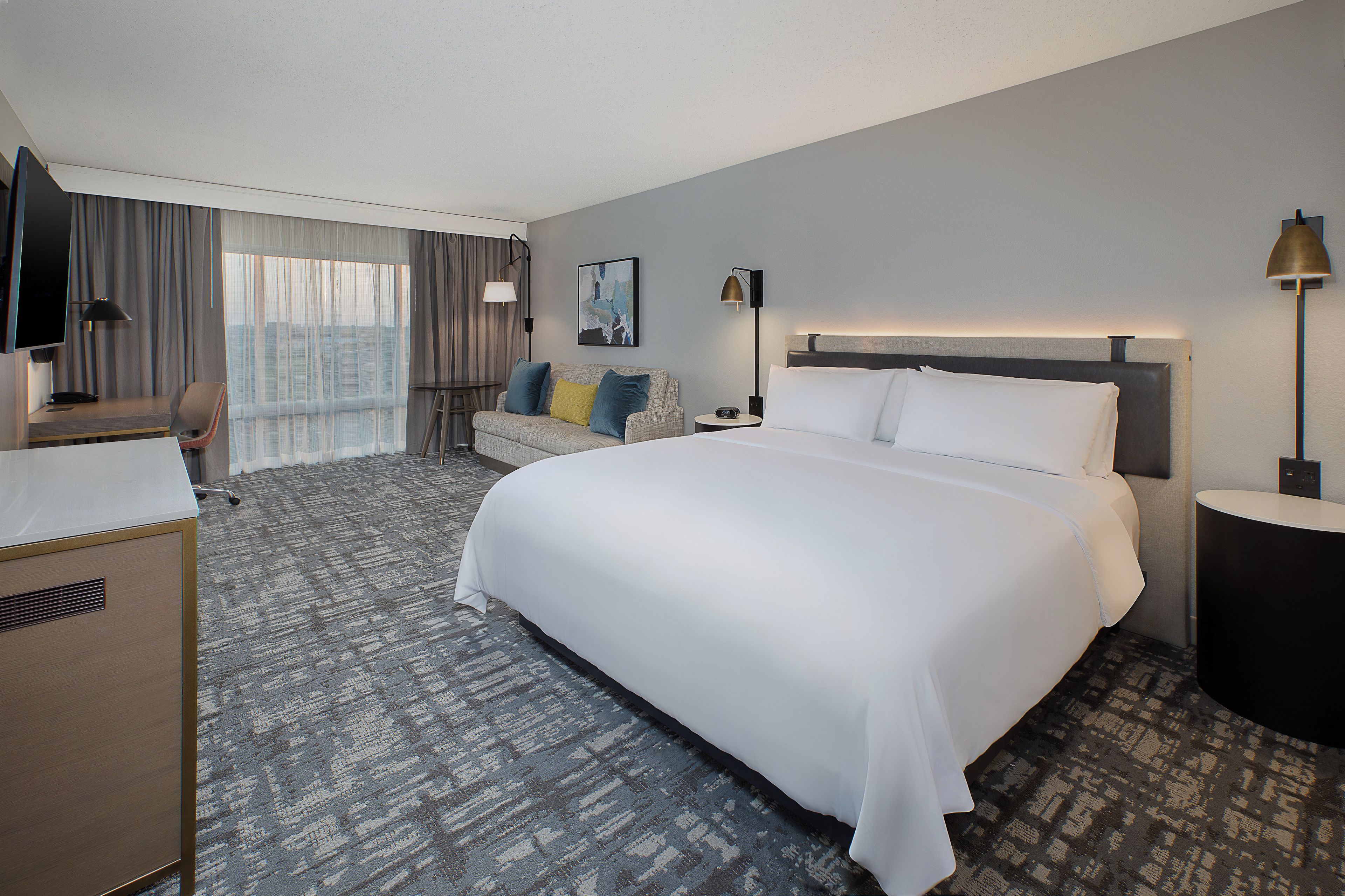 A hotel room with a large bed, a desk, a chair, a TV, and a window with curtains.