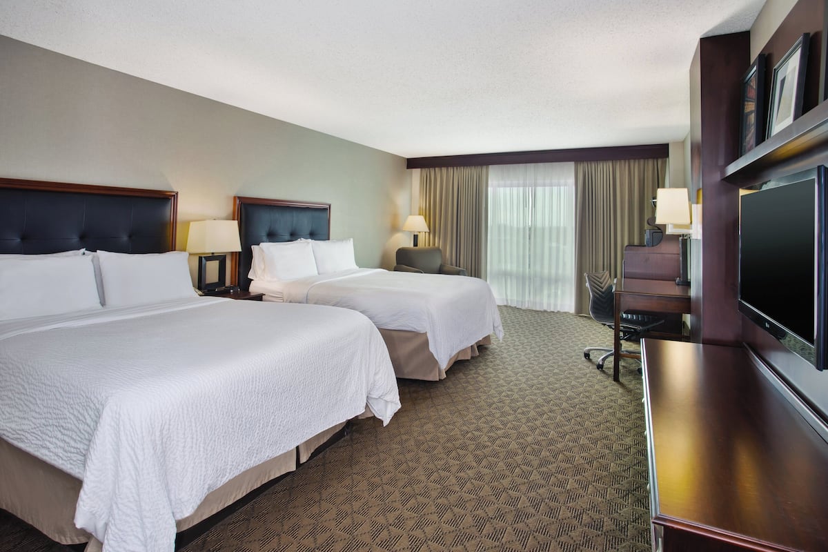 A hotel room with two beds, a desk, a TV, and a large window with curtains.