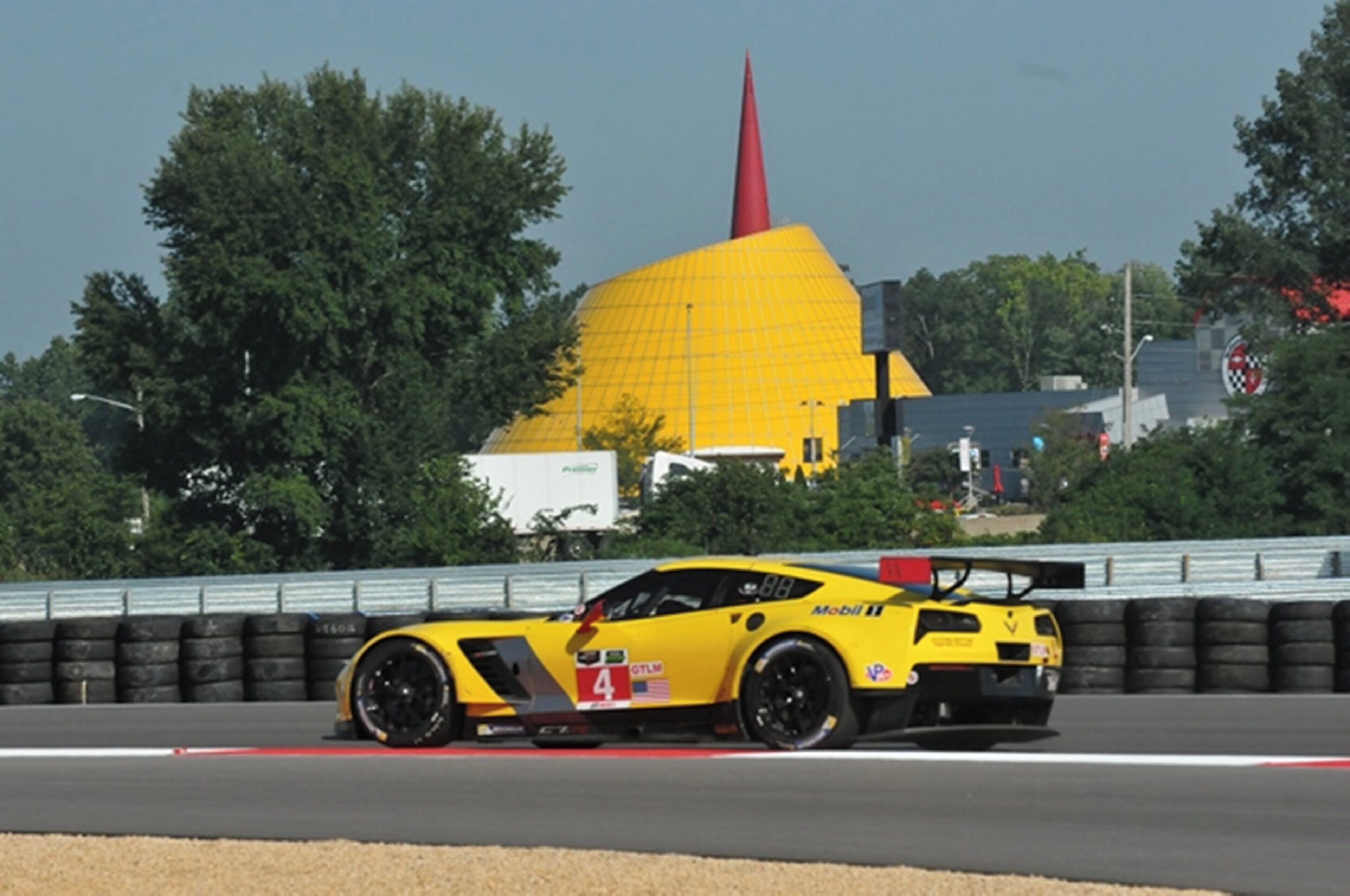 A yellow sports car on a race track with a large yellow building in the background.