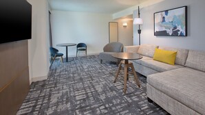 A modern hotel room with a large flat-screen TV, a grey sofa with a yellow pillow, a round coffee table, a blue chair, and a patterned carpet. - Crowne Plaza Bowling Green KY by IHG (Bowling Green)