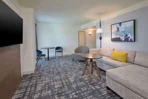 A modern hotel room with a large flat-screen TV, a grey sofa with a yellow pillow, a round coffee table, a blue chair, and a patterned carpet. - Crowne Plaza Bowling Green KY by IHG (Bowling Green)
