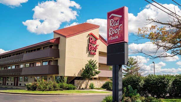 Un Red Roof Inn con techo rojo y paredes beige, un estacionamiento y vegetación circundante.