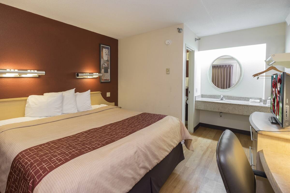 A hotel room with a large bed, a desk with a chair, a round mirror, and a small window with curtains.