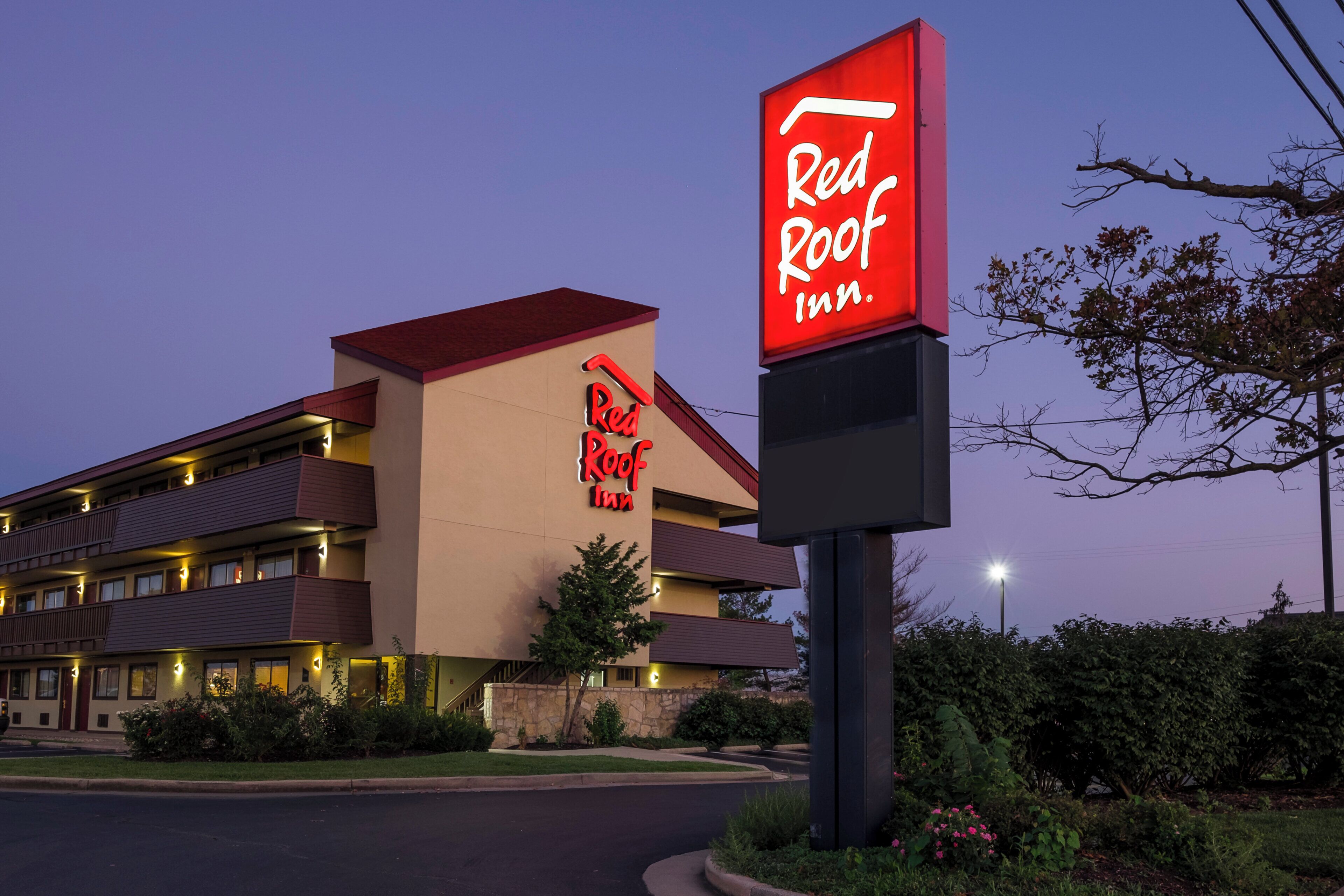 A Red Roof Inn with a modern design, featuring a prominent red and white sign, multiple balconies, and a well-lit entrance.