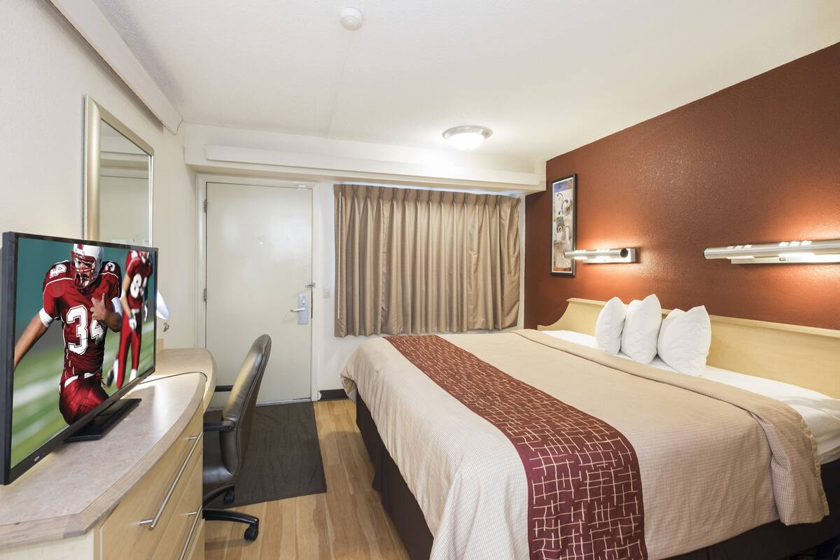 A hotel room with a bed, a desk with a TV, a chair, a door, and a window with curtains.