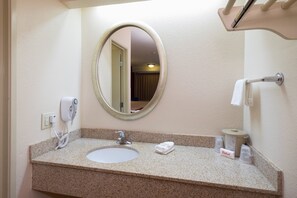 A hotel bathroom with a granite countertop, a round mirror, a sink, a towel dispenser, and a toilet paper holder. - Red Roof Inn Cincinnati - Sharonville (Sharonville)