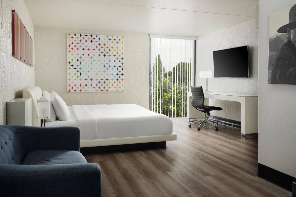 A hotel room with a bed, a desk, a chair, a TV, and a large window with blinds.