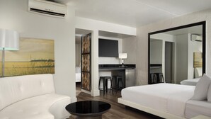 A modern hotel room with a white sofa, a round black coffee table, a bed with white bedding, a flat-screen TV mounted above a desk, and a large mirror.