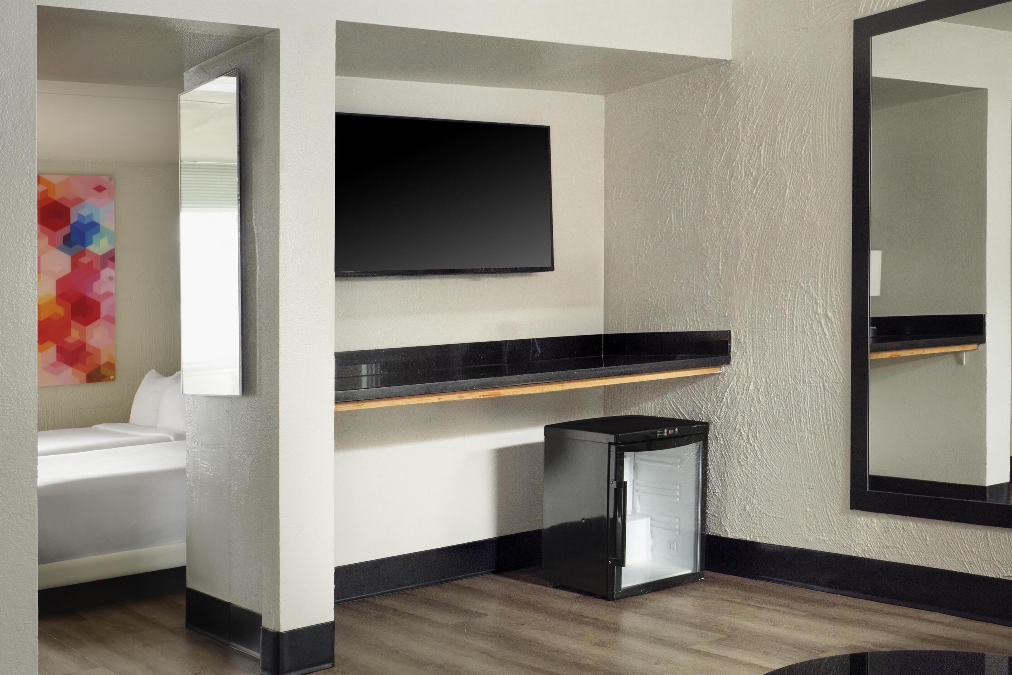 A modern hotel room with a bed, a flat-screen TV, a small refrigerator, and a large mirror.
