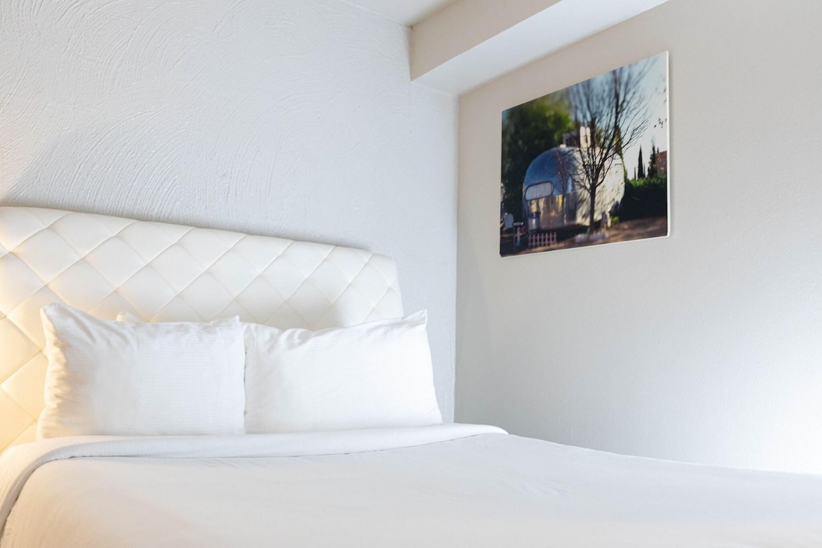 A hotel room with a white bed, a quilted headboard, and a framed picture on the wall.
