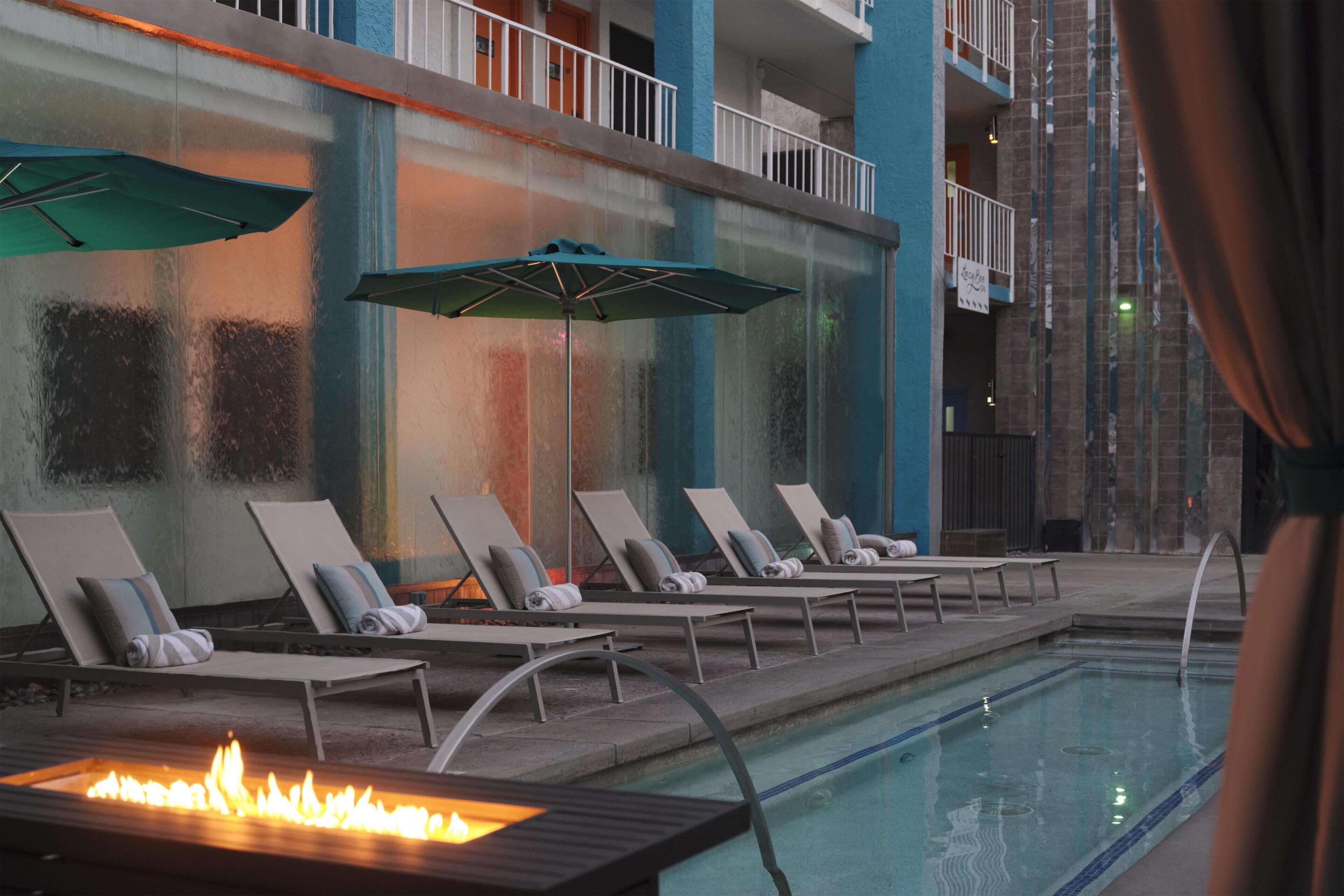 A poolside area with lounge chairs and umbrellas, a fire feature, and a view of the building's exterior.