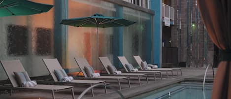 A poolside area with lounge chairs and umbrellas, a fire feature, and a view of the building's exterior.