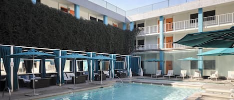 A swimming pool with lounge chairs and umbrellas, a multi-story building with balconies, and a hedge.