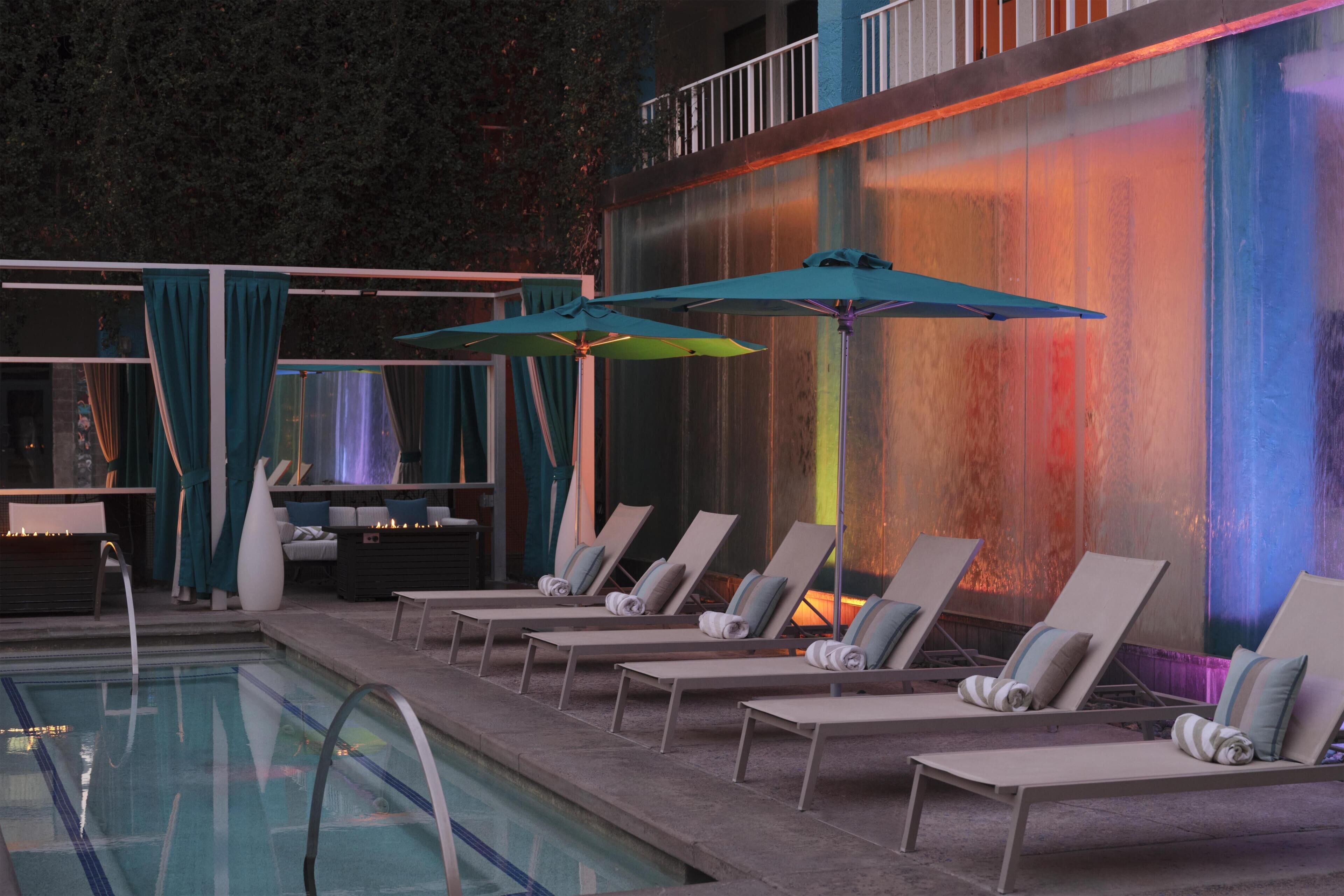 Un espace aménagé au bord de la piscine, avec des chaises longues et des parasols, une piscine, et un mur agrémenté d’un jeu de lumière coloré.