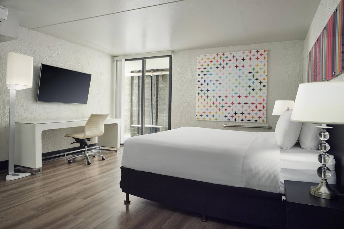 A hotel room with a bed, desk, chair, TV, and artwork on the wall.