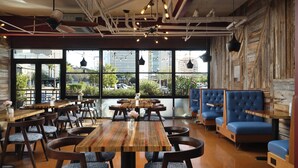 A restaurant with wooden tables and blue upholstered chairs, large windows with a view of buildings and trees, and exposed ceiling pipes.
