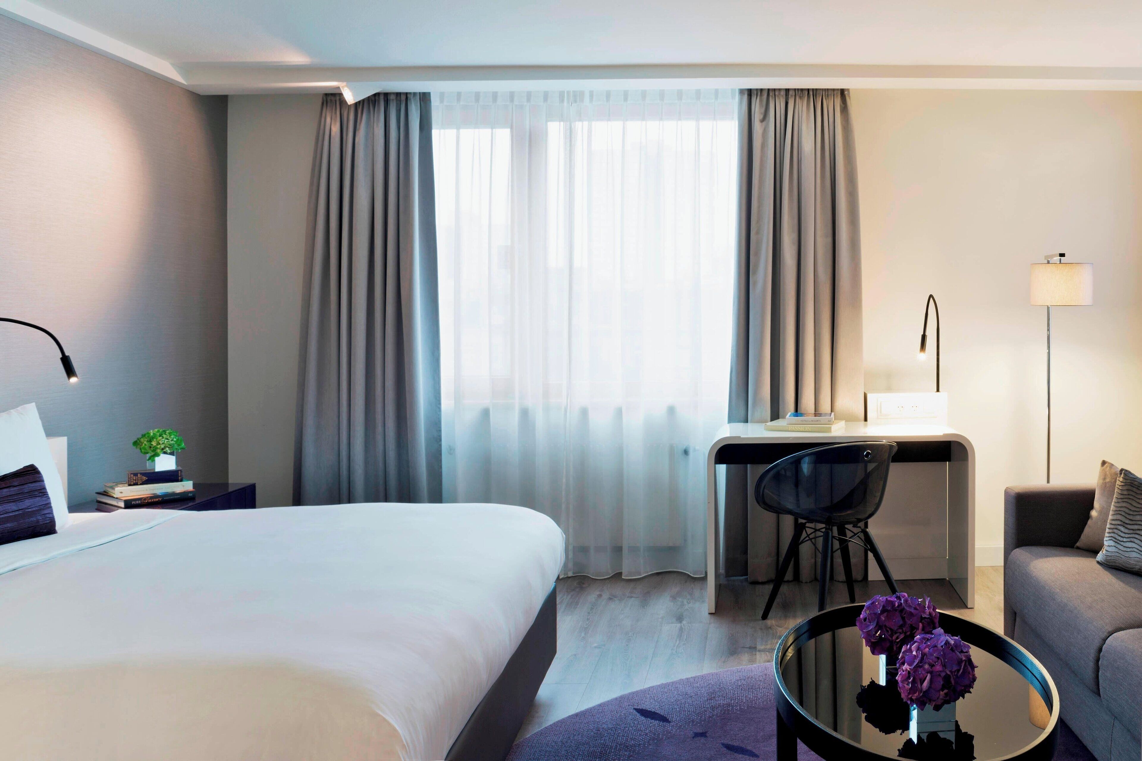 A hotel room with a bed, a desk, a chair, a sofa, and a coffee table with flowers.