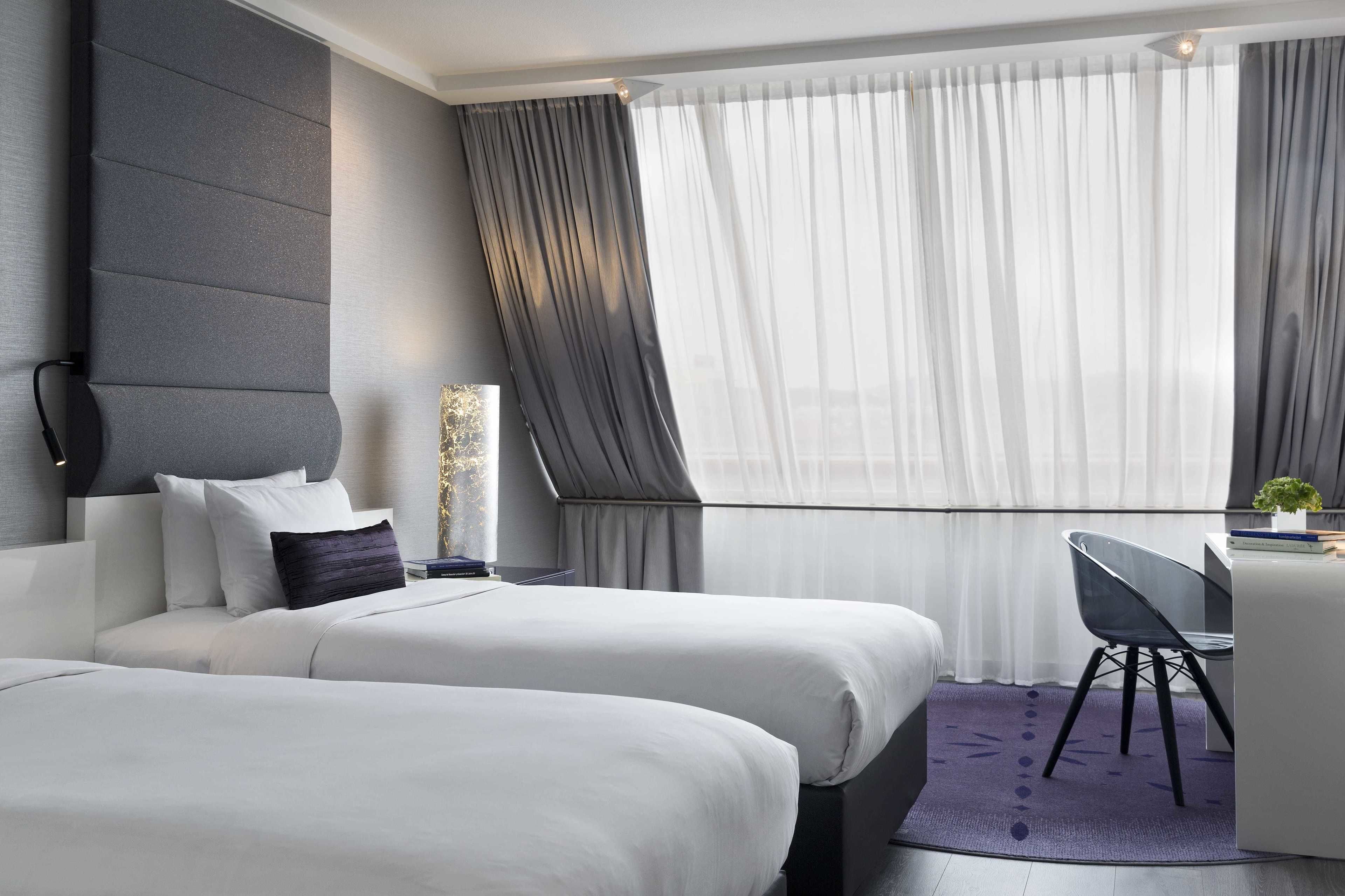 A modern hotel room with two beds, a desk, and a large window with sheer curtains.