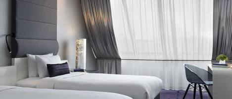 A modern hotel room with two beds, a desk, and a large window with sheer curtains.
