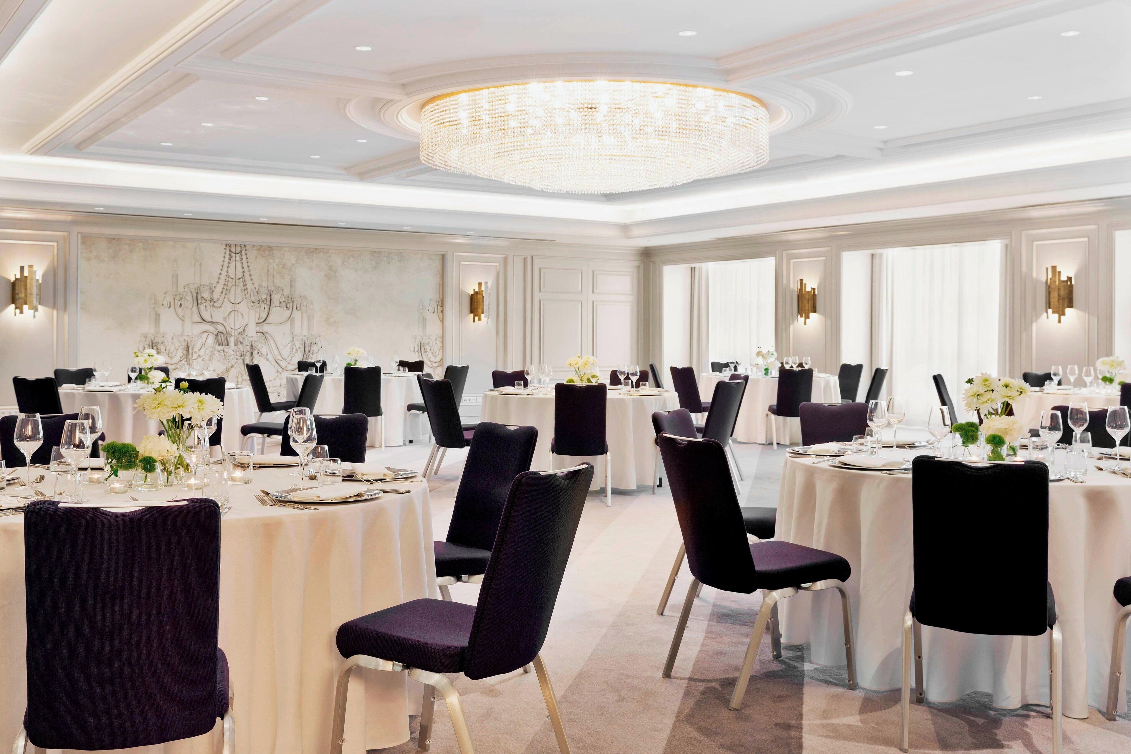 A banquet hall with round tables, black chairs, and chandeliers.