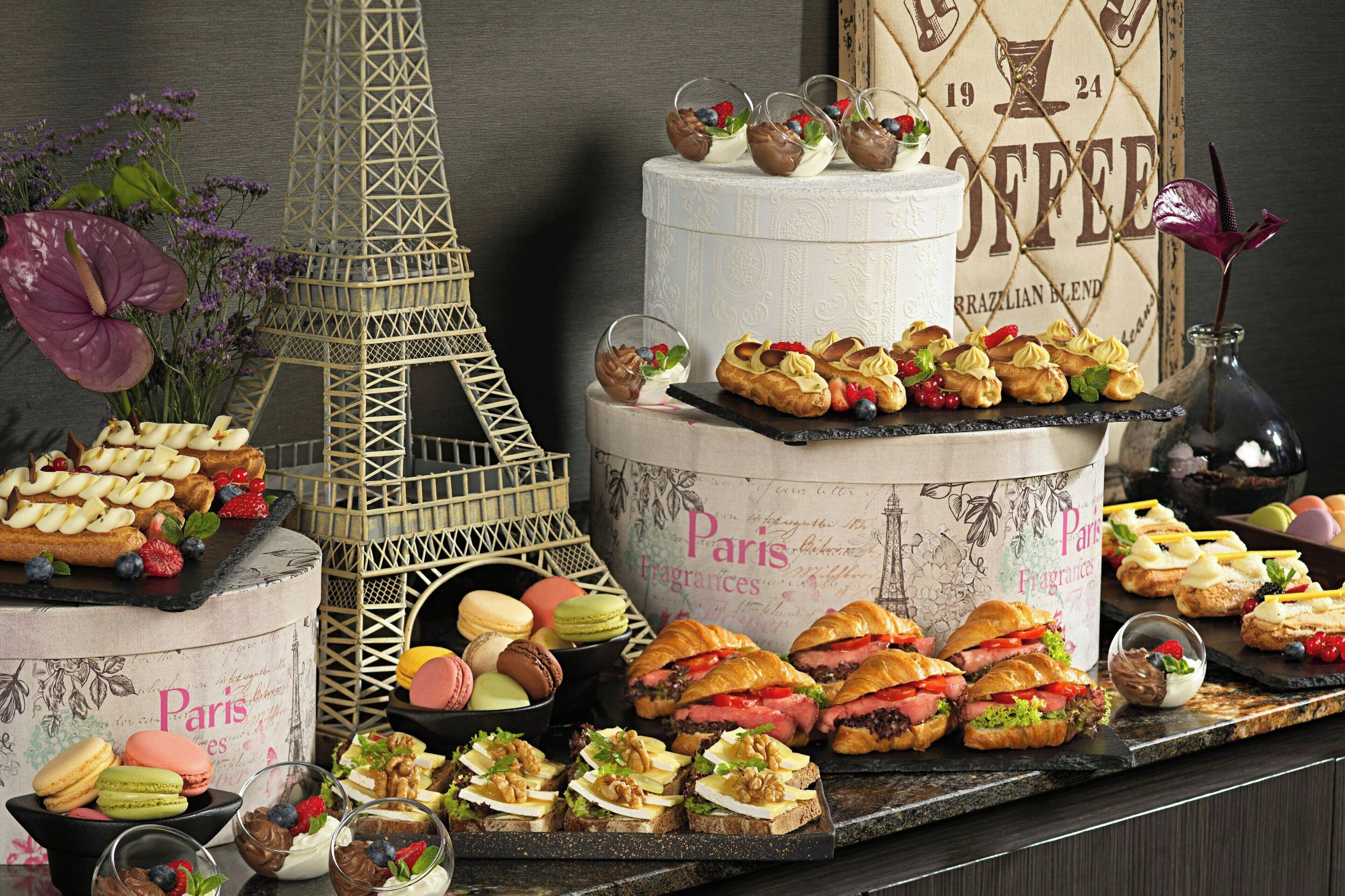 A variety of pastries and desserts displayed on a tiered stand, with a decorative Eiffel Tower and a vintage coffee advertisement in the background.