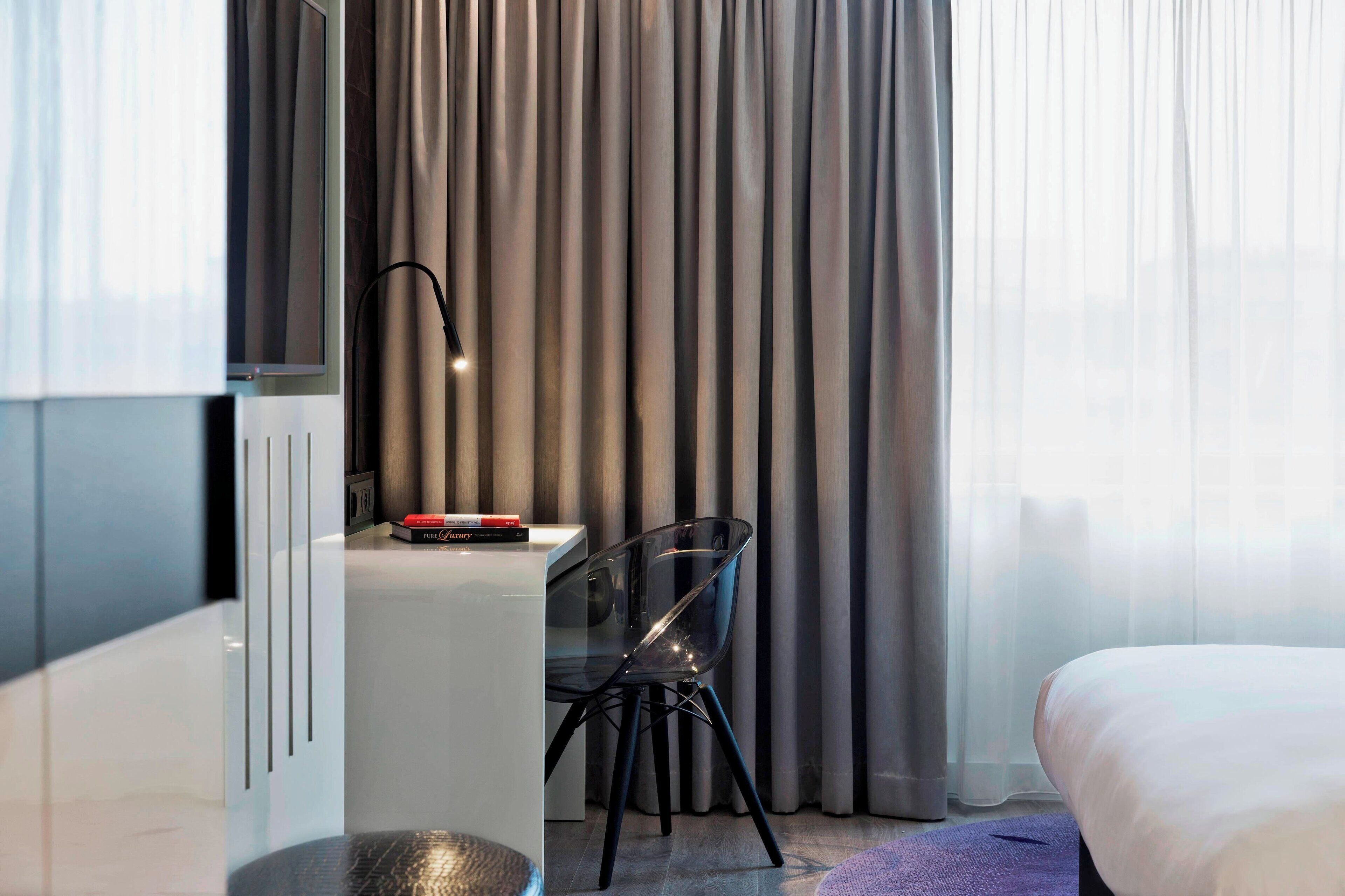 Una habitación de hotel moderna con cama, escritorio con silla, televisión y ventana con cortinas.