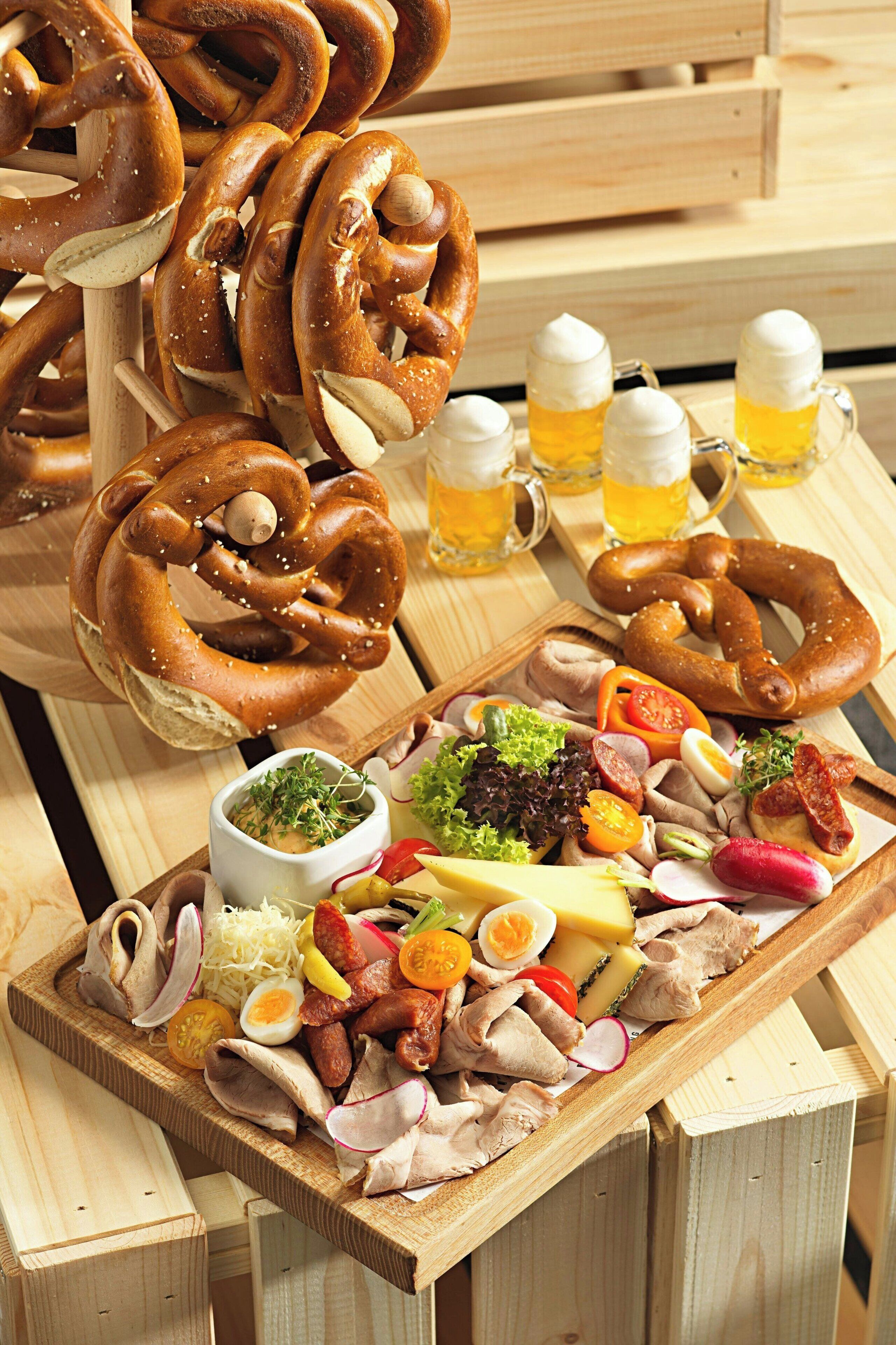 A wooden table with pretzels, a charcuterie board with meats, cheese, and vegetables, and beer mugs.