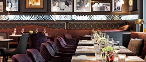 A restaurant with velvet chairs, tables set with wine glasses and menus, and framed pictures on the wall.