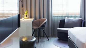 Habitación de hotel moderna con cama, escritorio y silla, un otomanillo redondo y vistas de la ciudad a través de amplias ventanas.