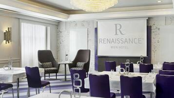 A conference room with a large screen displaying "Renaissance Wien Hotel". There are multiple tables with chairs, a chandelier, and a patterned wall.