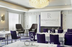 Una sala conferenze con un grande schermo che mostra "Renaissance Wien Hotel". Sono presenti diversi tavoli con sedie, un lampadario e una parete decorata.