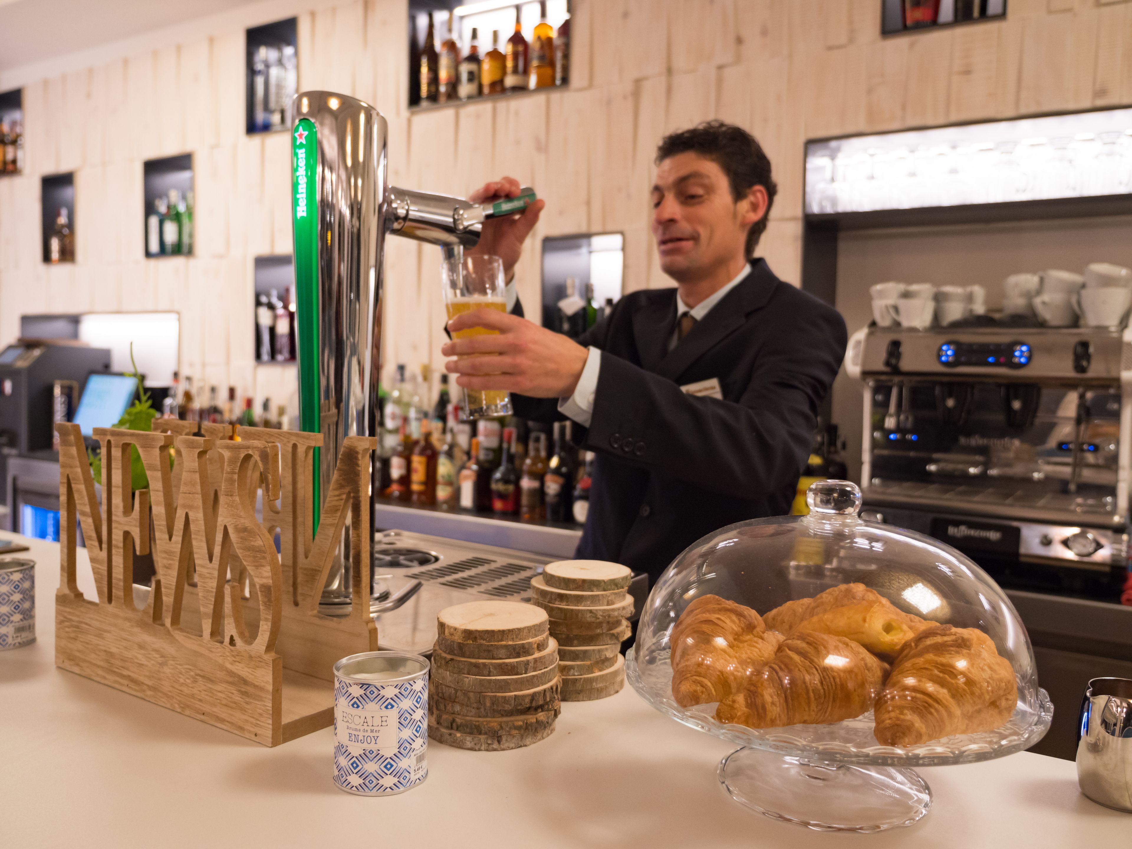Een barista schenkt een drankje in een glas, een presentatie van croissants en een biertappunt.