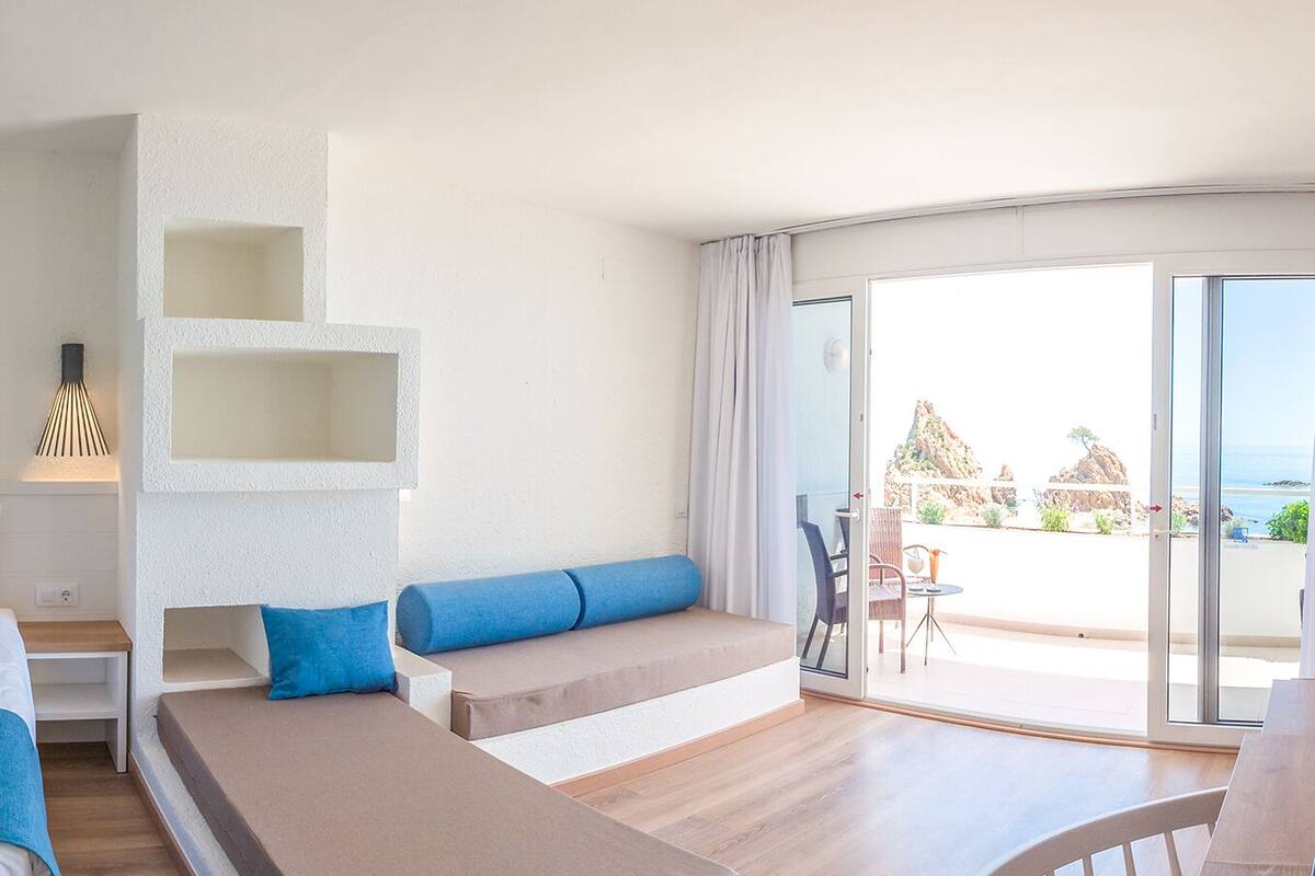 A modern hotel room with a bed, a seating area, a coffee station, and a view of the sea.