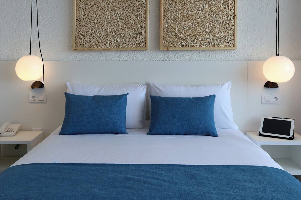A neatly made bed with two blue pillows and a white duvet, flanked by two bedside tables with lamps and a phone.