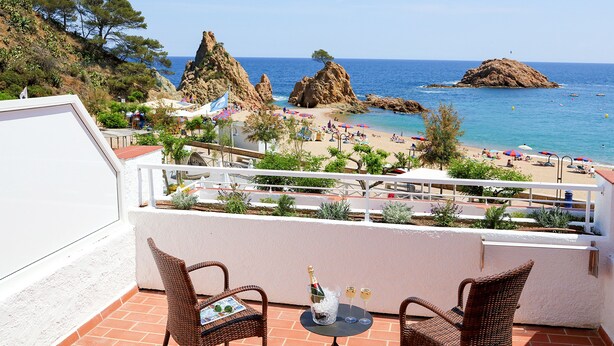 A terrace with wicker chairs, a table with a bottle and glasses, and a view of a beach with rocky formations and umbrellas. - Golden Mar Menuda (Tossa de Mar)